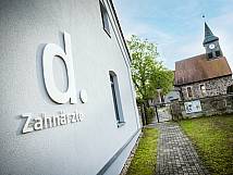 Praxisfassade mit Logo „d. Zahnärzte“ und Kirche im Hintergrund.